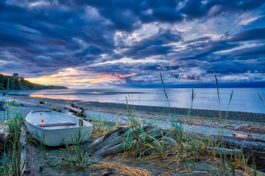 A derelict rowboat sits on a reedy shoreline, Kye Bay in the background. The sky is dark blue with colourful cloud formations.